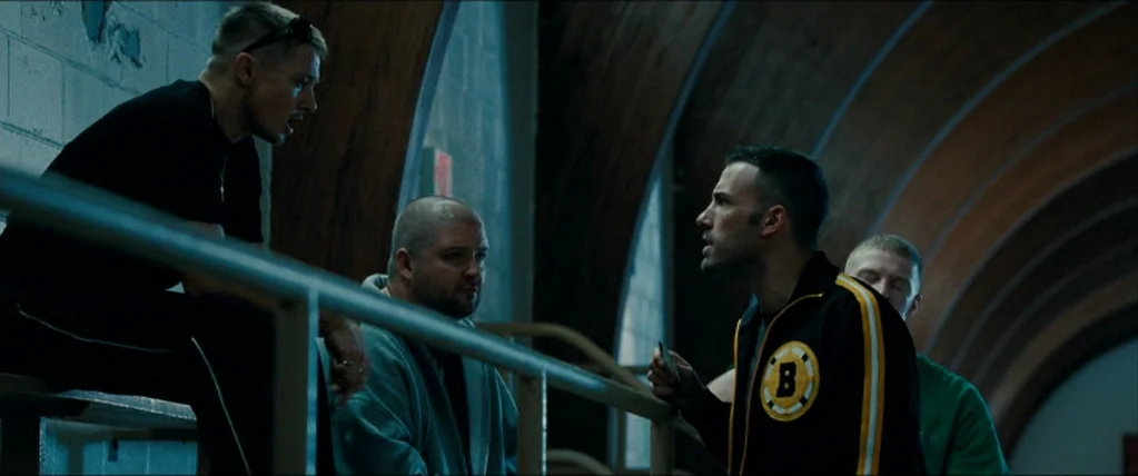 The Town (2010) movie still: medium — A group of men sit on high-arched gymnasium bleachers under a low ceiling. One man talks directly to another…