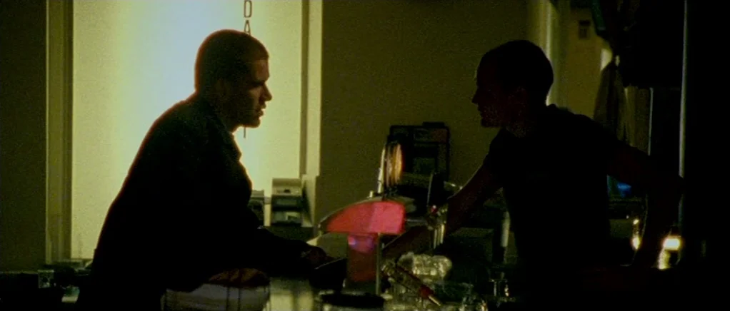 Reconstruction (2003) movie still: medium — A dark indoor space with a backlit doorway and a service area counter. Two men lean over a bar counter…