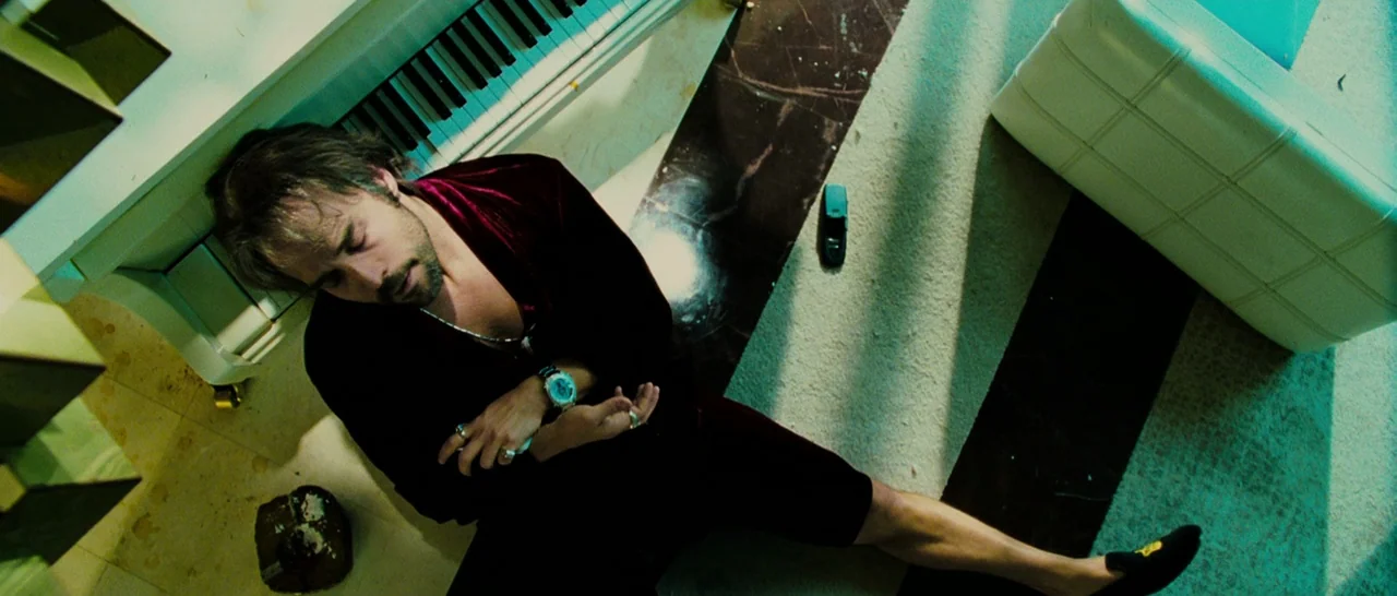 Slice (2018) movie still: medium — A high-angle view shows a man sitting on a marble and carpeted floor. He is slumped against a white piano next to…