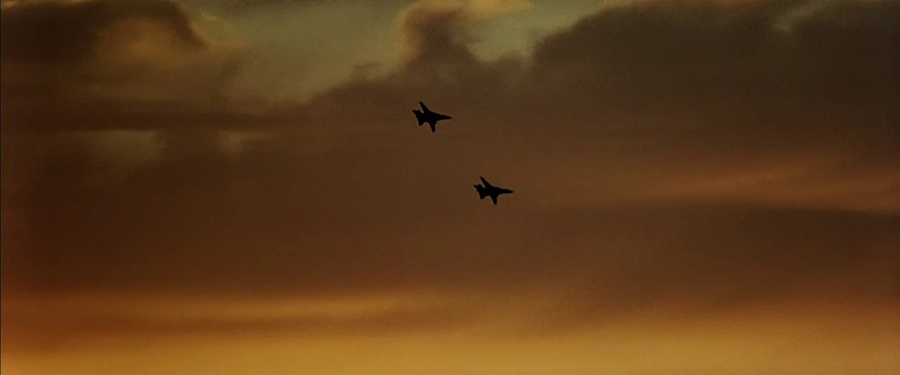 Top Gun (1986) movie still: wide — Two fighter jets fly through a golden-hued sky filled with scattered sunset clouds. The aircraft appear as dark…