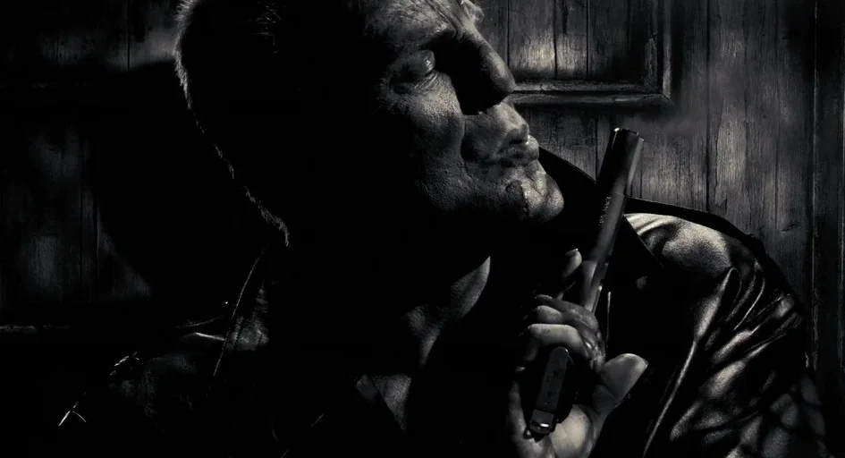 Sin City (2005) movie still: closeup — A dark space with vertical wood-paneled walls and sharp, directional lighting. A muscular man in a leather…