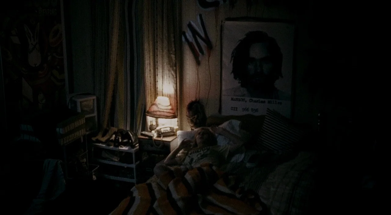 Halloween 2 (2009) movie still: wide — A dimly lit room with vintage 1970s decor and posters on the walls. A young woman lies in bed, illuminated by…
