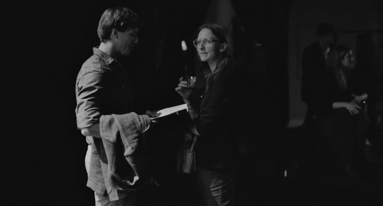 Frances Ha (2012) movie still: medium — A dimly lit indoor gathering with dark surroundings and blurred figures in the distance. A man and woman…