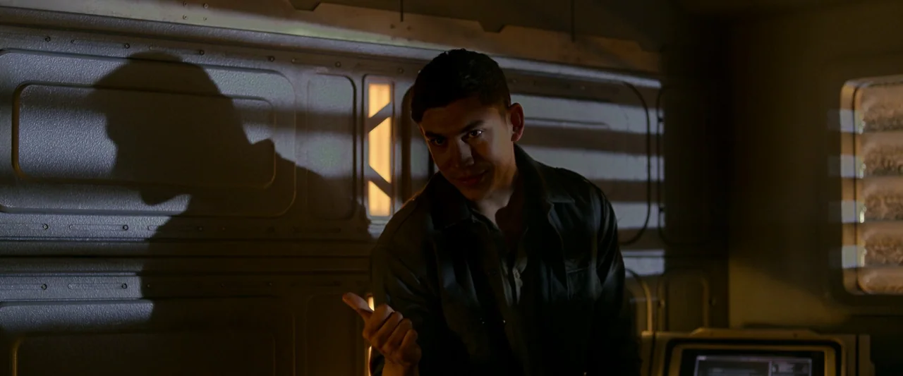 Alien: Romulus (2024) movie still: medium — A dimly lit room with modular metal wall paneling and a glowing control interface. A man in a dark shirt…