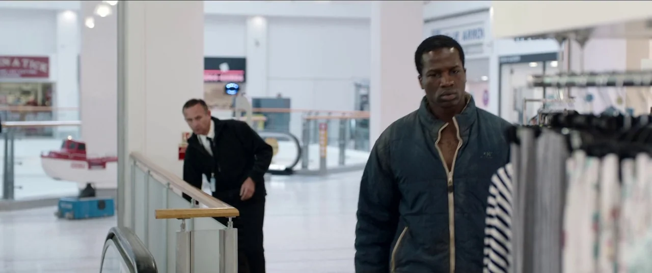 His House (2020) movie still: medium — A brightly lit interior of a multi-level shopping center with white tile floors. A man walks in the…
