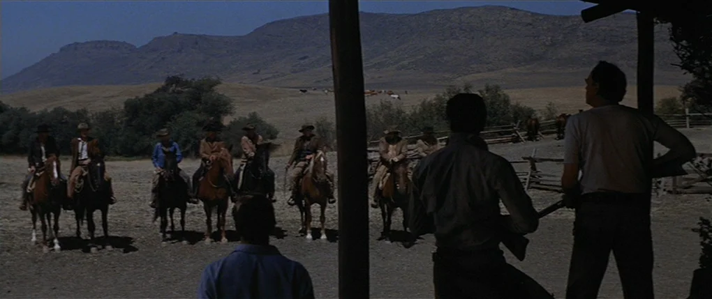 Flaming Star (1960) movie still: wide — A rural ranch yard with dusty ground sits beneath a range of desert hills. A group of men on horses faces…