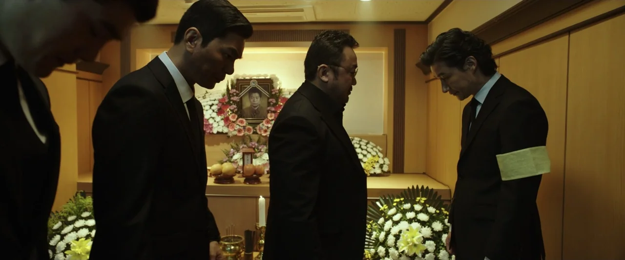 The Gangster, the Cop, the Devil (2019) movie still: medium — A funeral altar with flowers and a portrait is located in a wood-paneled room. Four…