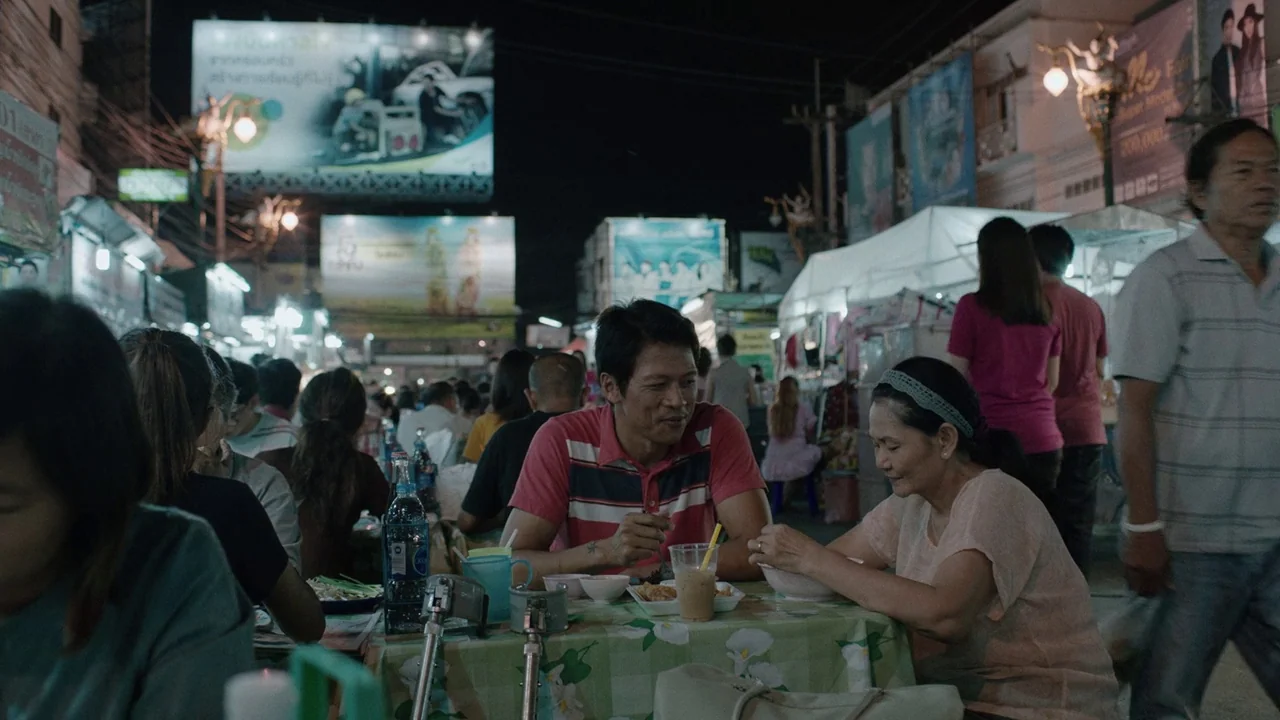Cemetery of Splendour (2015) movie still: medium — A bustling outdoor night market with illuminated billboards and crowded pedestrian stalls. A man…