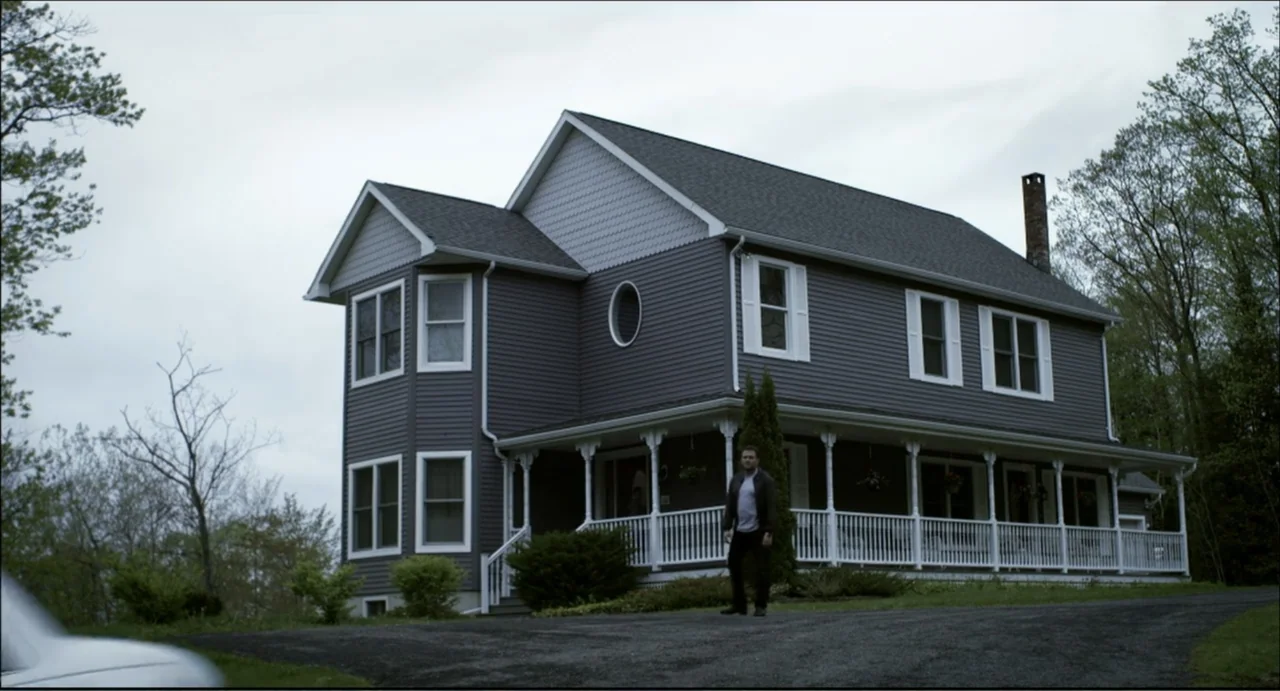 The Secret Village (2013) movie still: wide — A two-story grey siding house with a white wrap-around porch stands under overcast skies. A man stands…