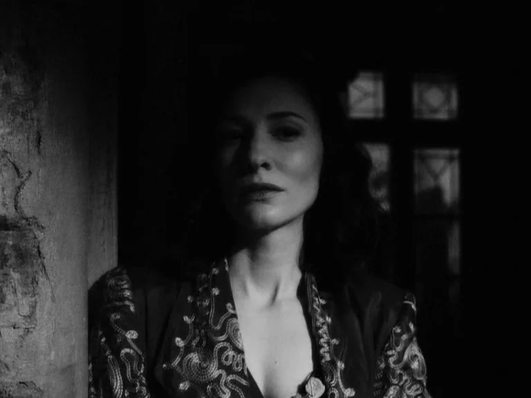 The Good German movie still: closeup — A dark indoor space features a woman standing against a rough textured wall. She wears an ornate embroidered…
