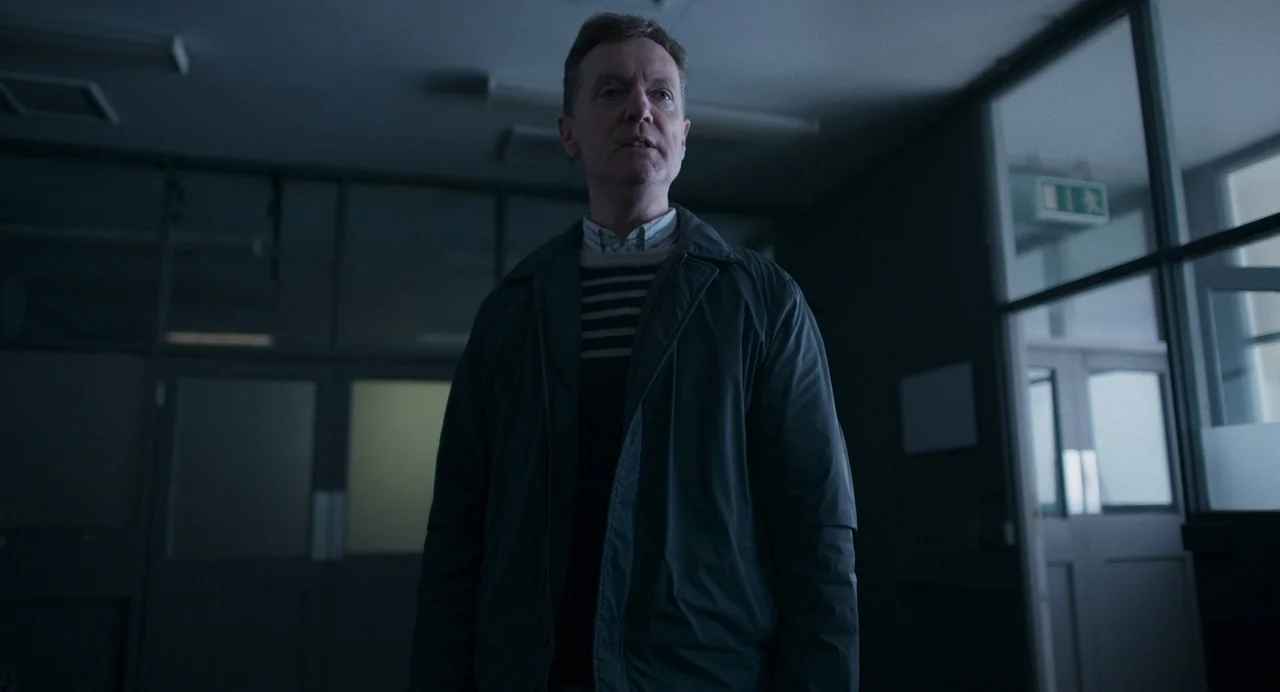 All You Need Is Death (2023) movie still: medium — A dimly lit office corridor features glass partitions and a green exit sign. A middle-aged man…