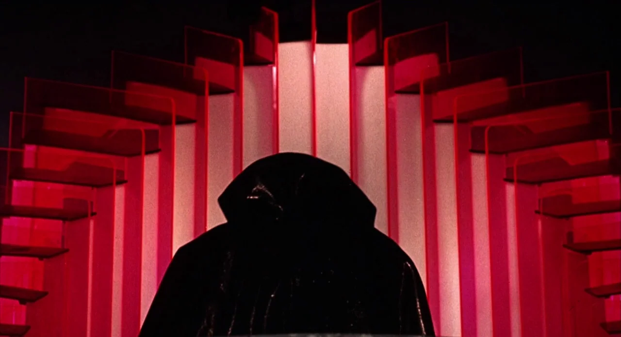 The Abominible Dr. Phibes (1972) movie still: medium — A dark, stylized stage features tiered red acrylic shelves radiating outward behind a…