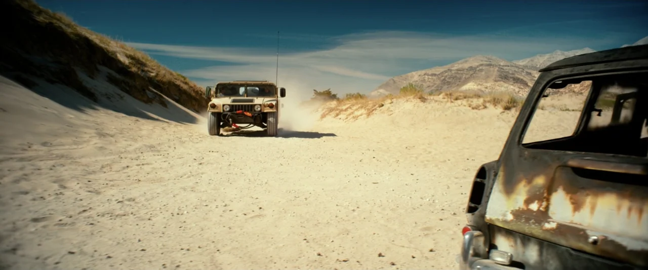 God Bless the Broken Road (2018) movie still: wide — A dry desert landscape features sand dunes and distant mountains under a blue sky. A tan…