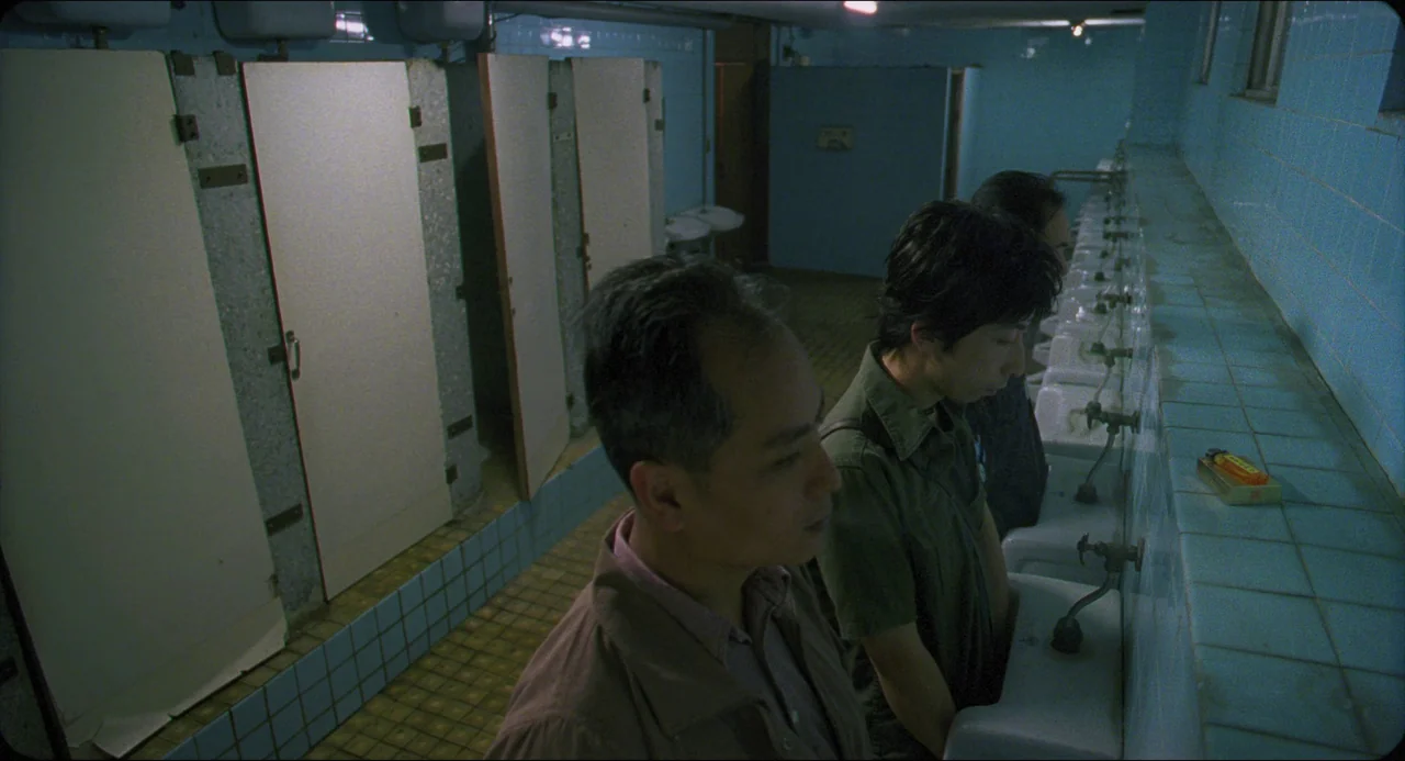 Goodbye, Dragon Inn (2003) movie still: medium — A public tiled restroom features a row of urinals against a cool blue wall. Two men stand at the…