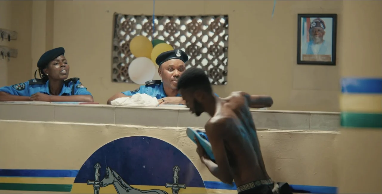 Project Assault (2025) movie still: medium — A Nigerian police station reception area features a desk and a framed portrait on wall. Two uniformed…