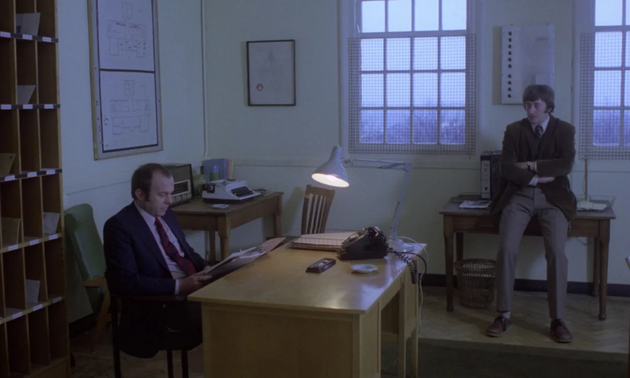 Scum (1979) movie still: wide — A quiet administrative room contains wooden desks, filing systems, and metal-gridded windows. Two men sit in the…