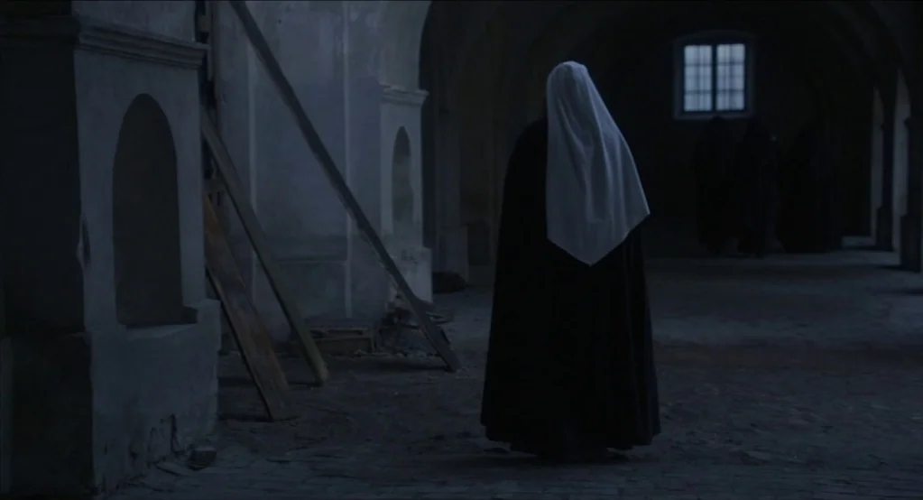 The Innocents (2016) movie still: fullbody — A dark, unfinished stone corridor with arched ceiling and wooden support beams. A solitary figure in a…