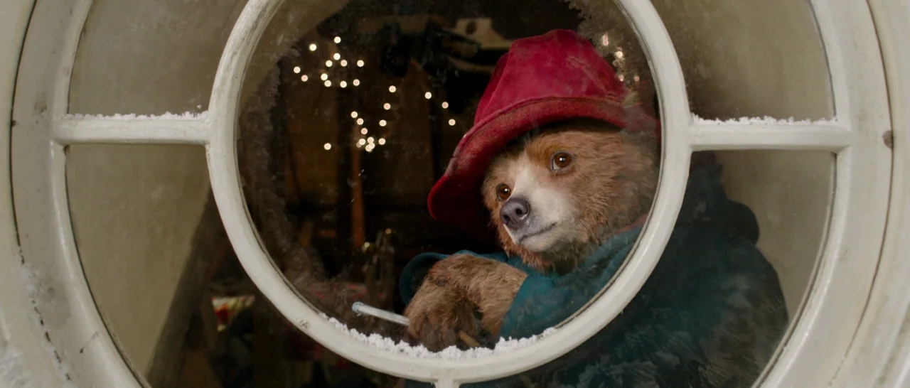 Paddington (2014) movie still: closeup — A brown bear looks through a circular glass windowpane framed by white wood trim. Small lights twinkle in…