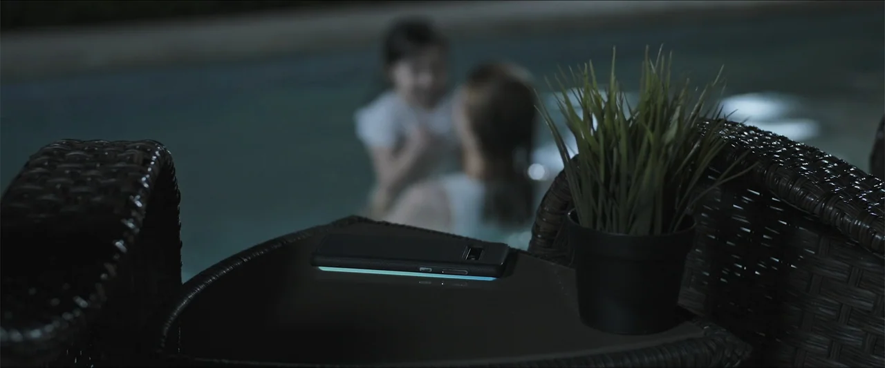 Los habitantes (2023) movie still: medium — A dark outdoor patio at night features a small potted plant and a smartphone. Two blurred figures are…