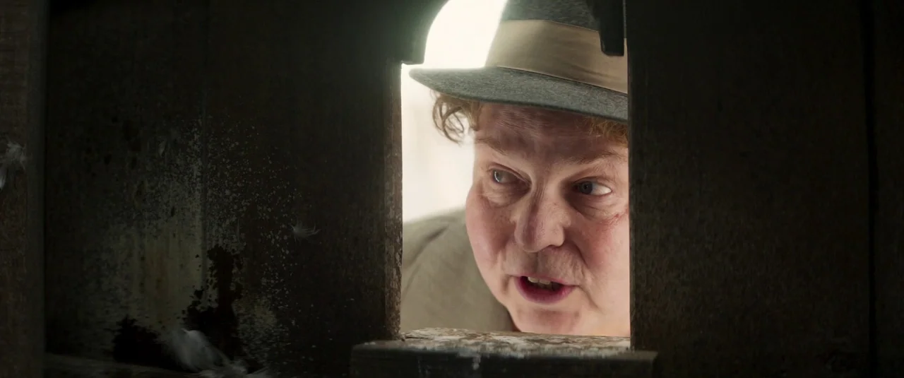 Wonka (2023) movie still: closeup — A man peers through a narrow rectangular slit in a dark wooden wall. Bright light from the outside highlights…