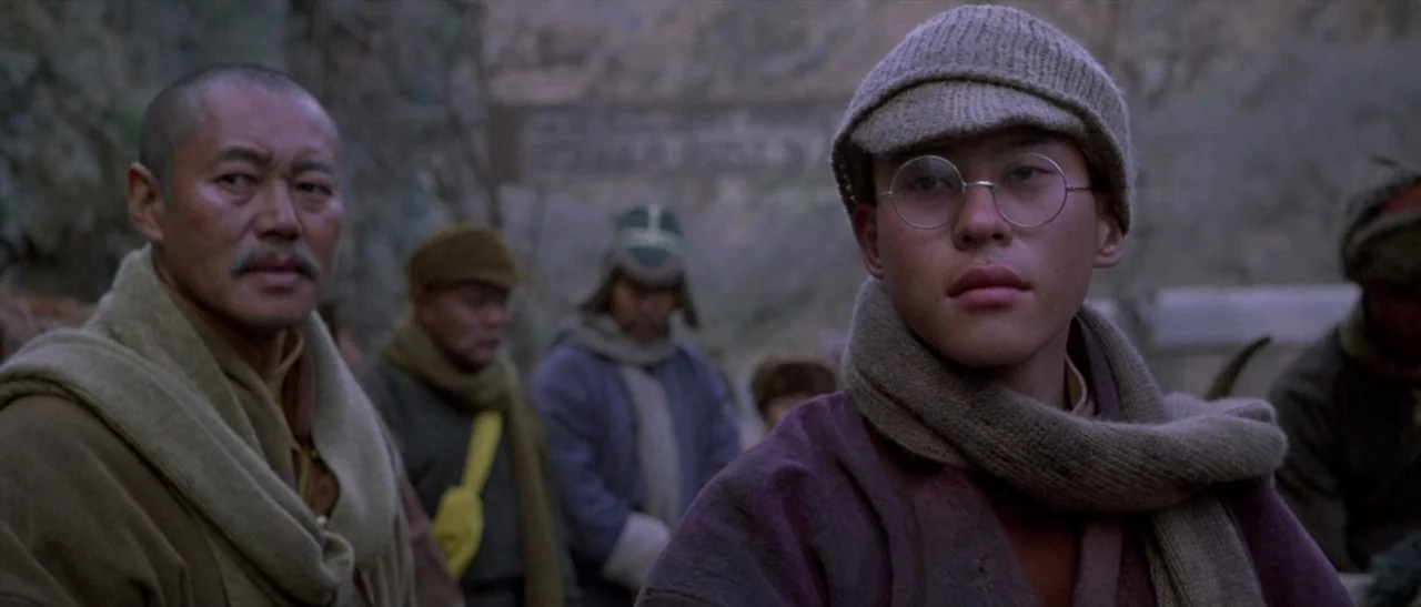 Kundun (1997) movie still: medium — A group of people standing on a rocky mountain path in cool daylight. A young man wearing glasses and a woolen…