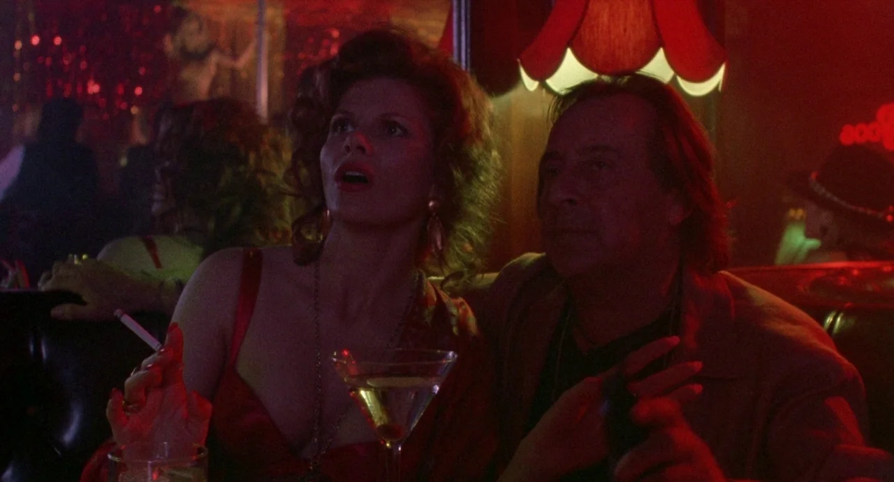 Touch (1997) movie still: medium — A dark, red-lit nightclub booth features two individuals seated closely together. The woman holds a cigarette in…