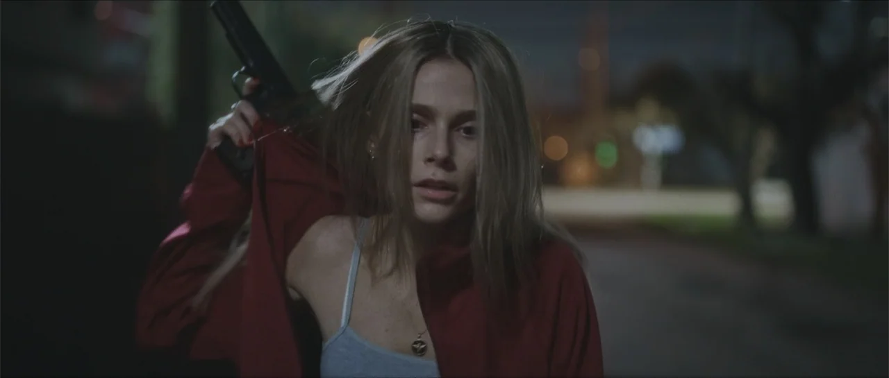 Nahir (2024) movie still: medium — A dark urban environment at night with blurred street lights in the background. A young woman in a red jacket…