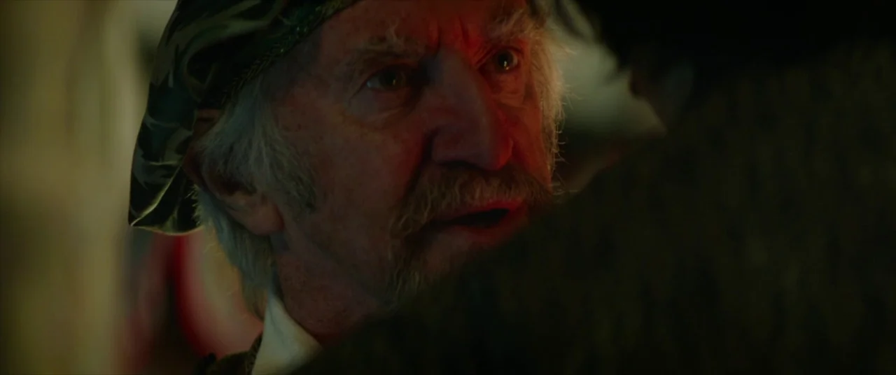 The Man Who Killed Don Quixote (2018) movie still: closeup — A dimly lit interior space features an older man wearing a textured cap. The man gazes…