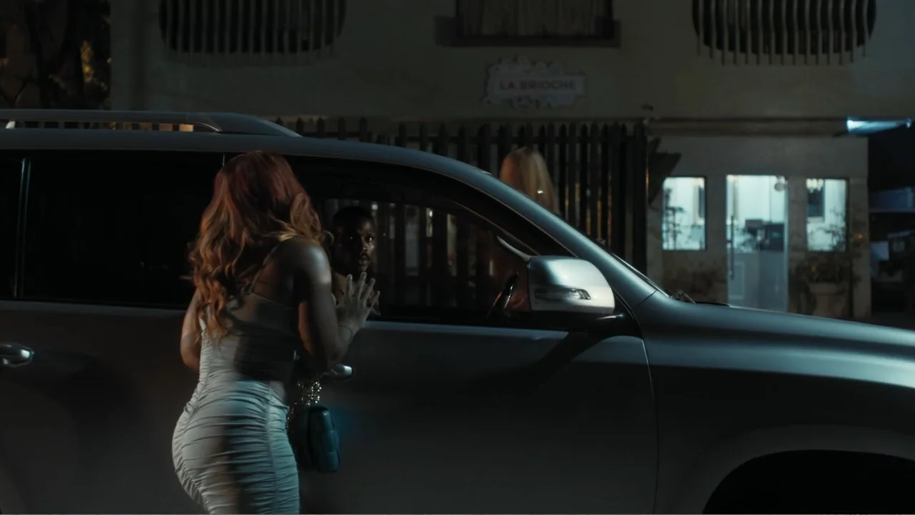 Gangs of Lagos (2023) movie still: medium — An exterior night setting features a silver SUV parked in a dark lot. A woman stands beside the driver's…