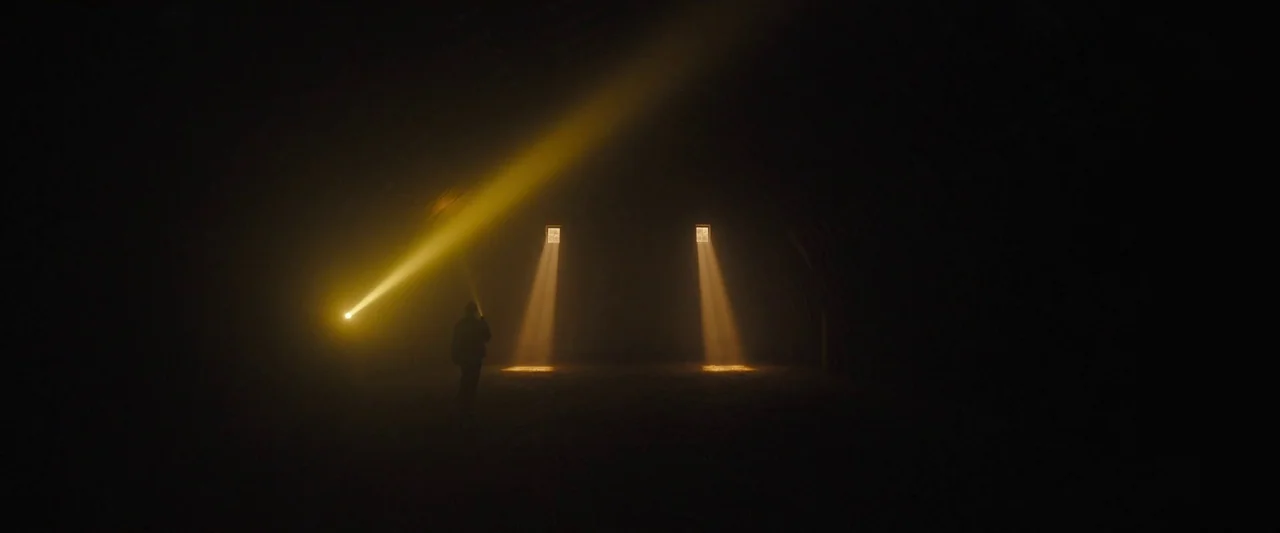 Longlegs (2024) movie still: wide — A vast, dark barn interior contains dust particles suspended in beams of light. A solitary silhouette stands…
