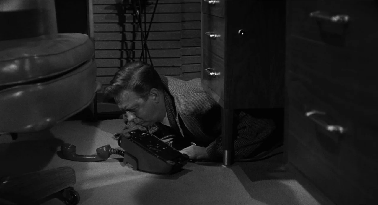The Fearmakers (1958) movie still: medium — A room features dark wood furniture and mid-century modern aesthetic details. A man crouches low to the…
