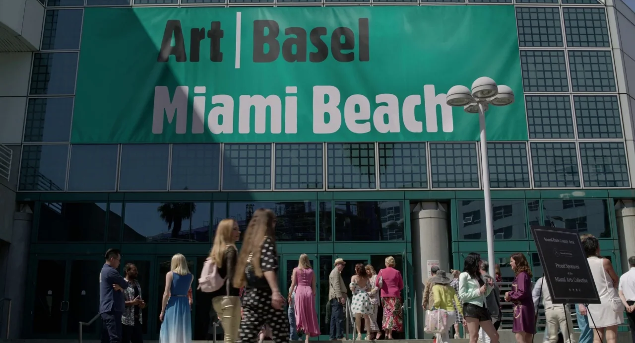 Velvet Buzzsaw (2019) movie still: wide — A modern glass and steel building displays a large green Art Basel Miami Beach banner. A diverse group of…