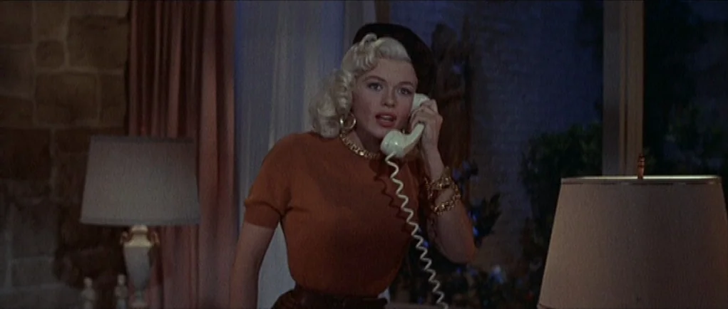 The Girl Can’t Help It (1956) movie still: medium — A dimly lit room features a stone fireplace wall and soft-shaded table lamps. A blonde woman…