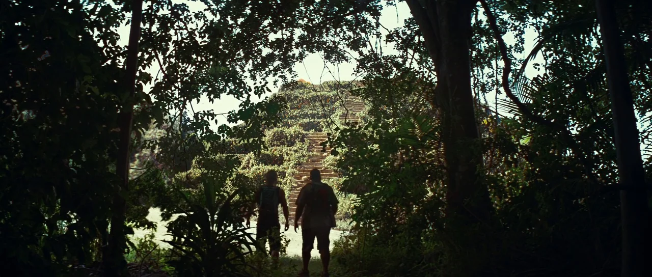 The Ruins (2008) movie still: wide — A dense green jungle surrounds an ancient overgrown stone pyramid with clearly visible steps. Two men are…
