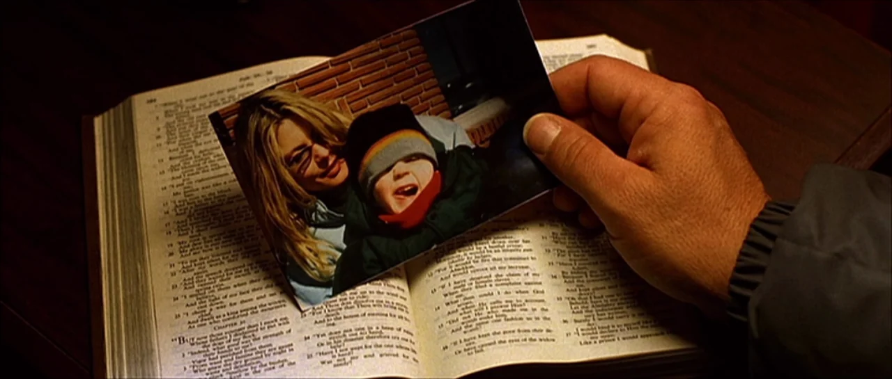 Fear X (2003) movie still: closeup — An open book with two columns of printed text rests on a dark surface. A hand holds a small photographic print…