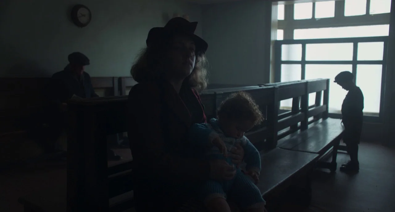 The Little Stranger (2018) movie still: medium — A dimly lit waiting area features wooden benches and a large paned window. A woman sits holding a…