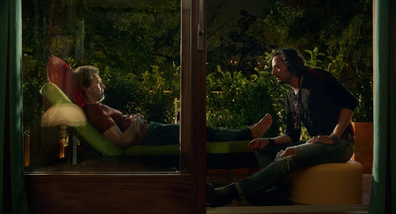 Pain and Glory (2019) movie still: wide — A nighttime balcony setting with lush green foliage surrounding two men on patio furniture. One man…
