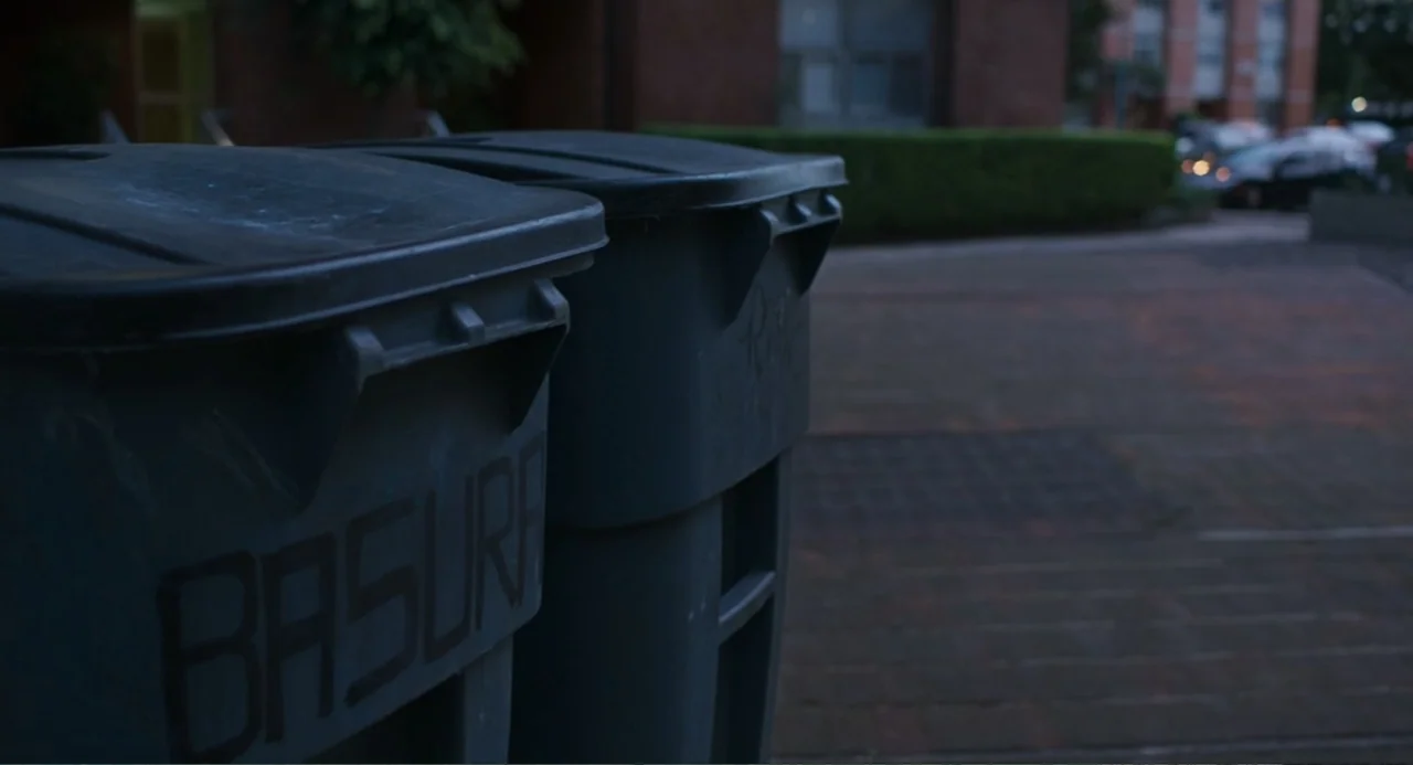 The Lift (2021) movie still: medium — Two plastic trash bins occupy the foreground in an empty paved outdoor space. Background includes a brick wall…
