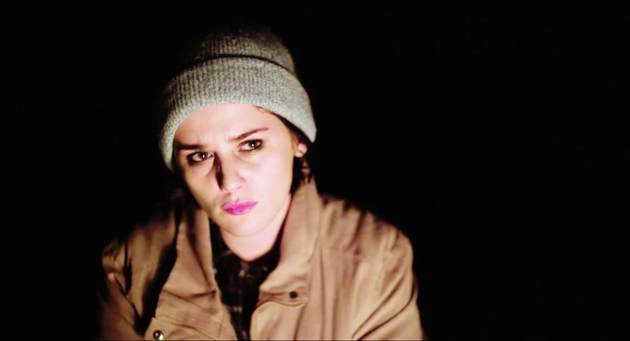 Like Me movie still: closeup — A dark outdoor space at night with a person lit by a fire. A woman in a beanie looks off-camera while bathed in low…