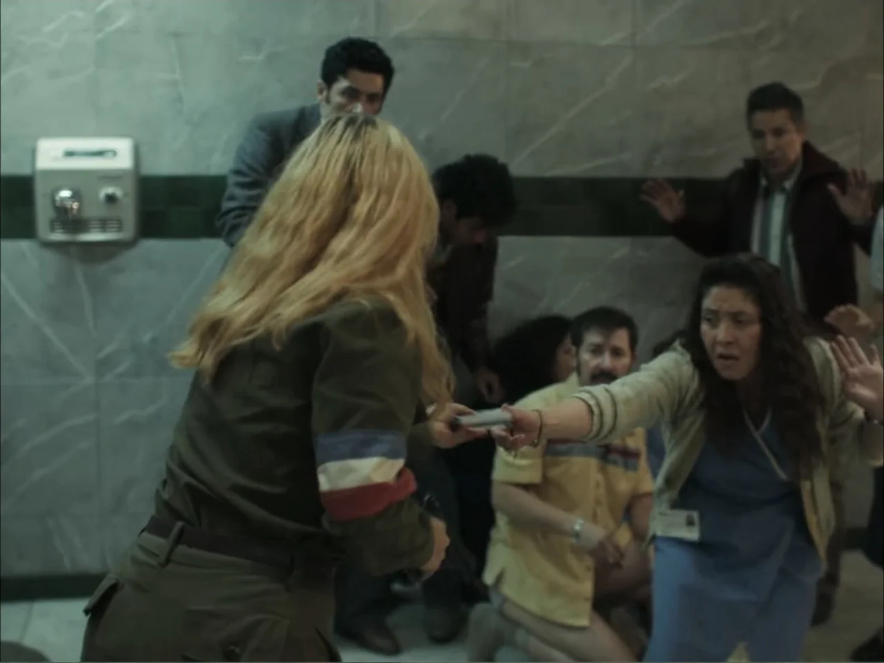 November (2025) movie still: medium — A utilitarian room with gray marble-tiled walls and a mounted hand dryer. A blonde woman exchanges a small…