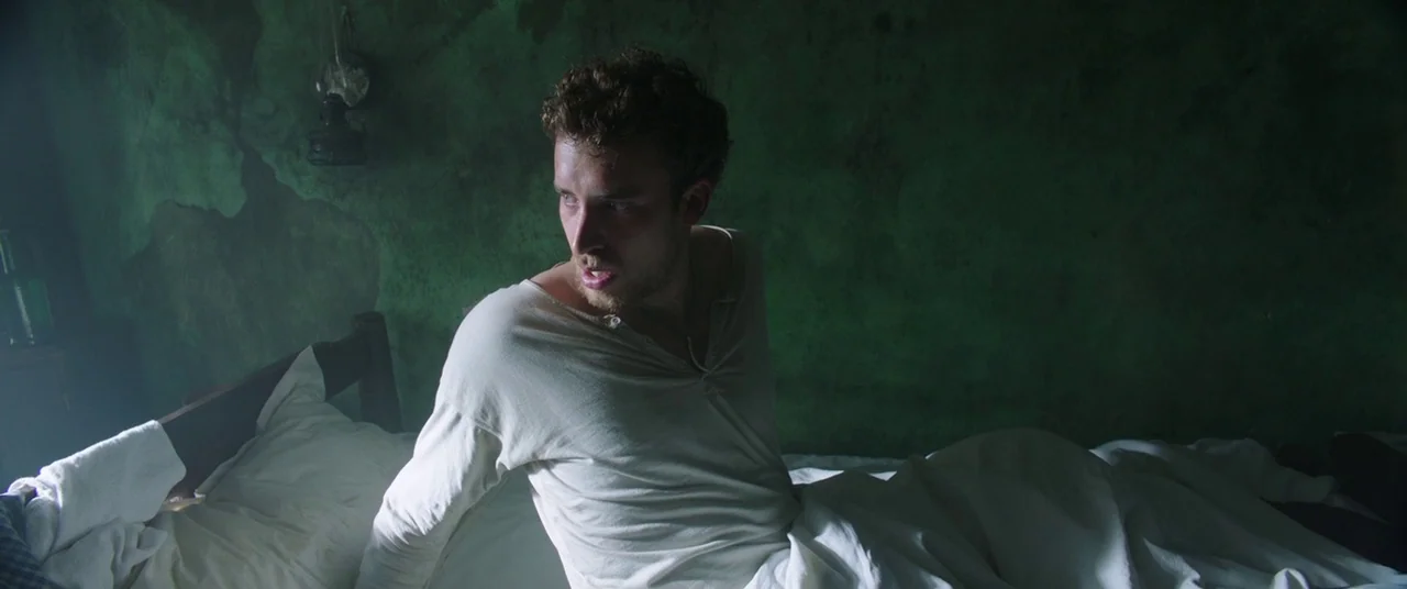 The Nun (2018) movie still: medium — A dimly lit room features a worn wall with peeling green paint. A man sits upright in bed while looking away…