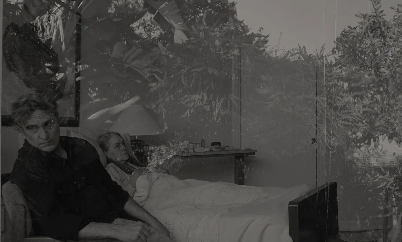 C’mon C’mon (2021) movie still: medium — A bedroom contains a man seated beside a woman in a hospital-style bed. Double-exposed foliage overlays the…