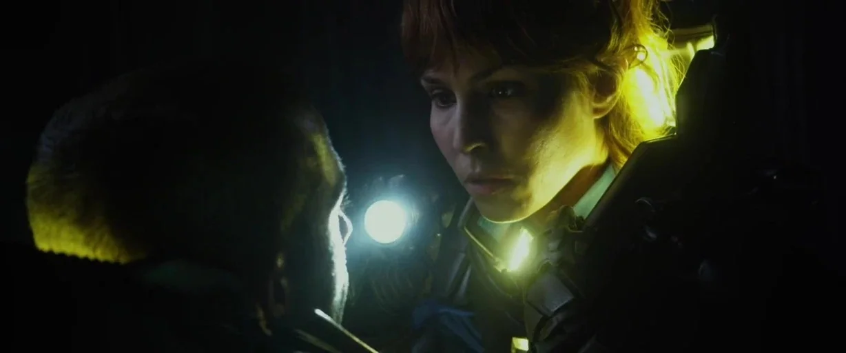 Prometheus (2012) movie still: medium — A dimly lit, metallic corridor features stark directional lighting on the subjects. A woman in tactical gear…