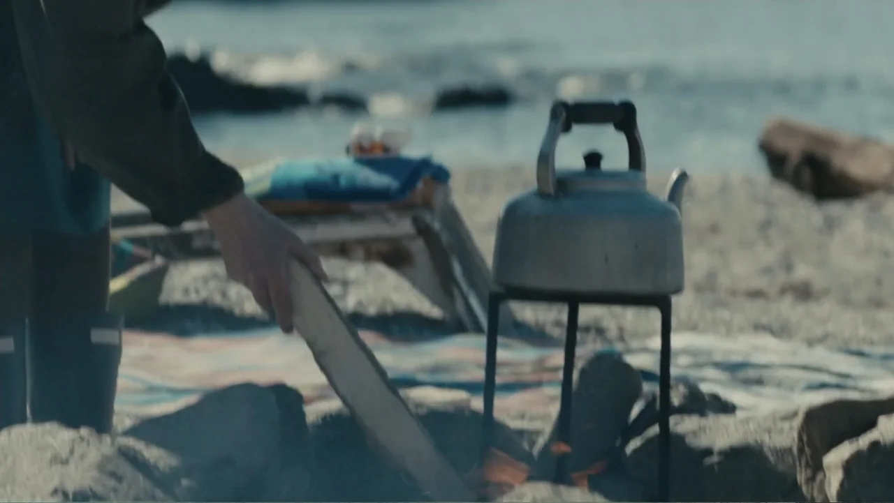 About Time (2013) movie still: medium — A pebble beach with a small campfire setup in the foreground near the water. A person in dark clothing…