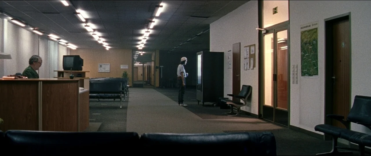 Luz (2018) movie still: wide — An expansive, sparsely furnished waiting area with rows of black vinyl chairs. A man sits at a desk while a woman…