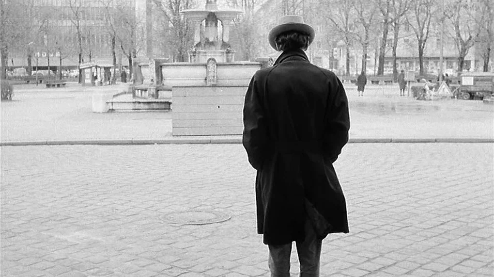 Love Is Colder Than Death (1969) movie still: fullbody — A stone-paved public square features a central tiered fountain surrounded by bare winter…