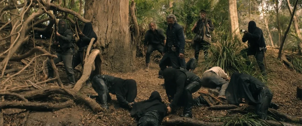 The Bluff (2026) movie still: fullbody — A muddy woodland clearing contains large tree roots and dense tropical foliage overhead. A group of armed…