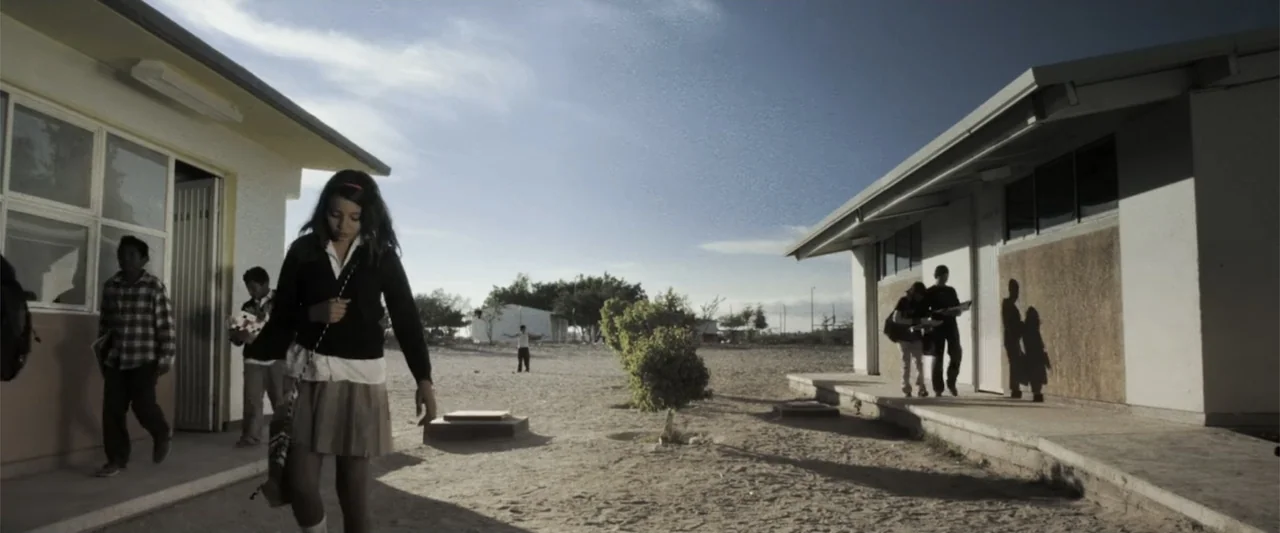 At Dawn (2013) movie still: fullbody — A sunny school courtyard features two parallel single-story buildings made of plain concrete blocks. A…