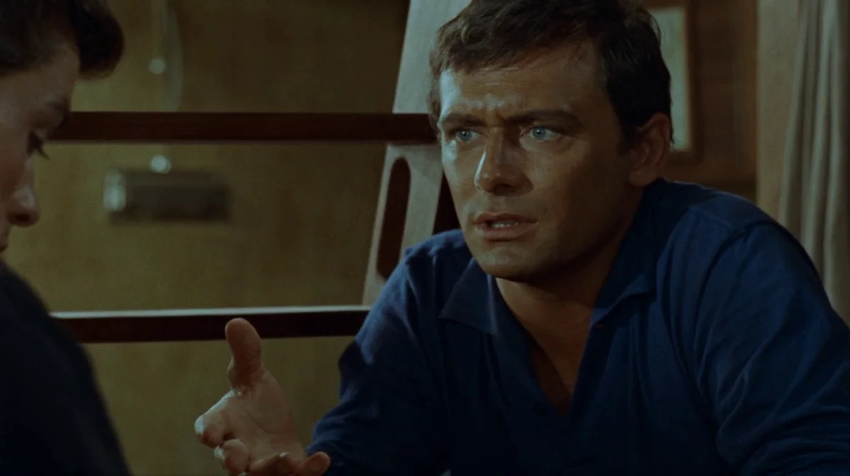 Purple Noon (Plein Soleil) (1960) movie still: medium — A man in a blue polo shirt gestures while sitting inside a rustic cabin. Wooden structural…