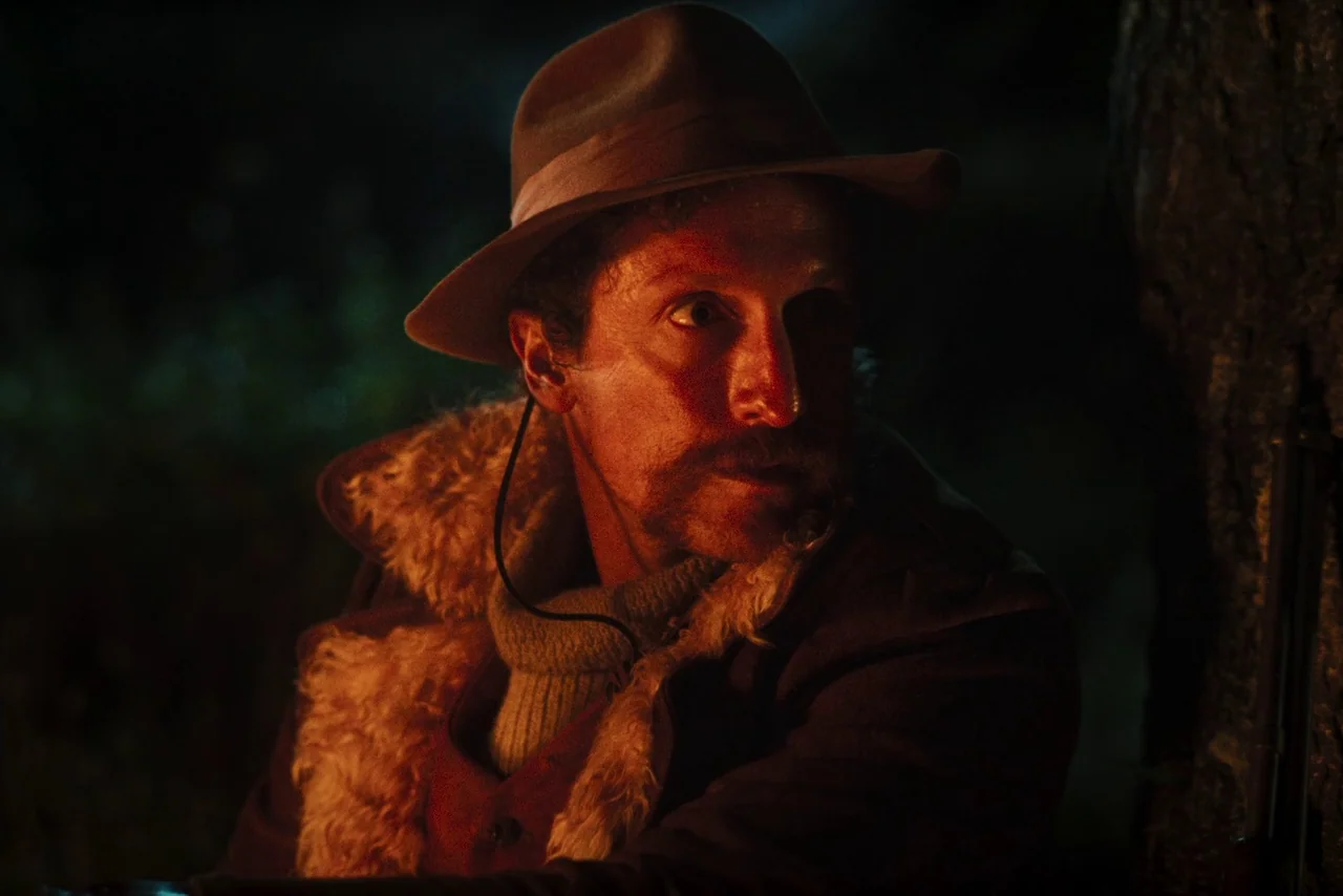 The Settlers (2023) movie still: closeup — A dark outdoor environment at night lit by a flickering fire. A man in a hat looks toward the side with…